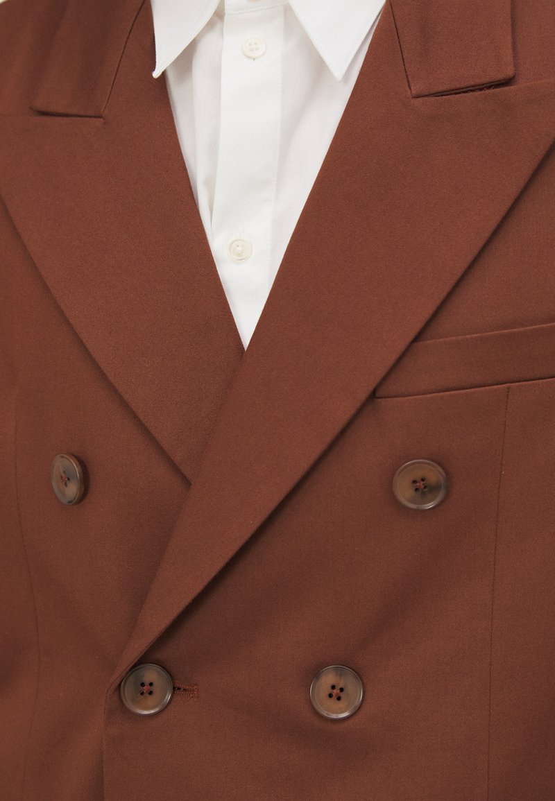 Blazer marrone doppiopetto realizzato in tessuto solido. Presenta un classico colletto a punte, due grandi bottoni marroni e una tasca sul petto.