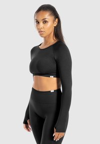 Kobieta o długich włosach, ubrana w czarny długi rękaw crop top i legginsy o wysokim stanie, stoi na tle jasnoszarym.