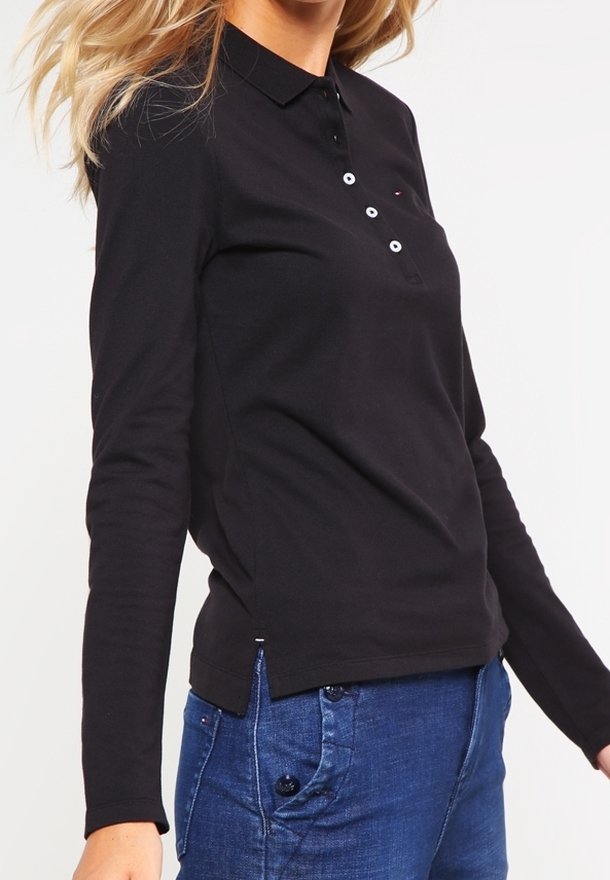 Schwarzes Langarm-Polohemd mit Kragen, ausgestattet mit drei weißen Knöpfen und einem seitlichen Schlitz. Kombiniert mit blauen Jeans.