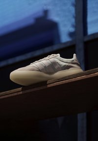 Ljusbeige sneaker i mocka och syntetmaterial, med tre vita ränder, rund tå och en genomskinlig sula.