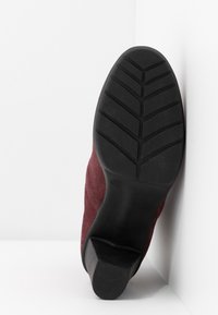 Semelle d'une chaussure à talon rouge foncé avec semelle en caoutchouc noir, positionnée à la verticale contre un mur et un sol blanc en angle.
