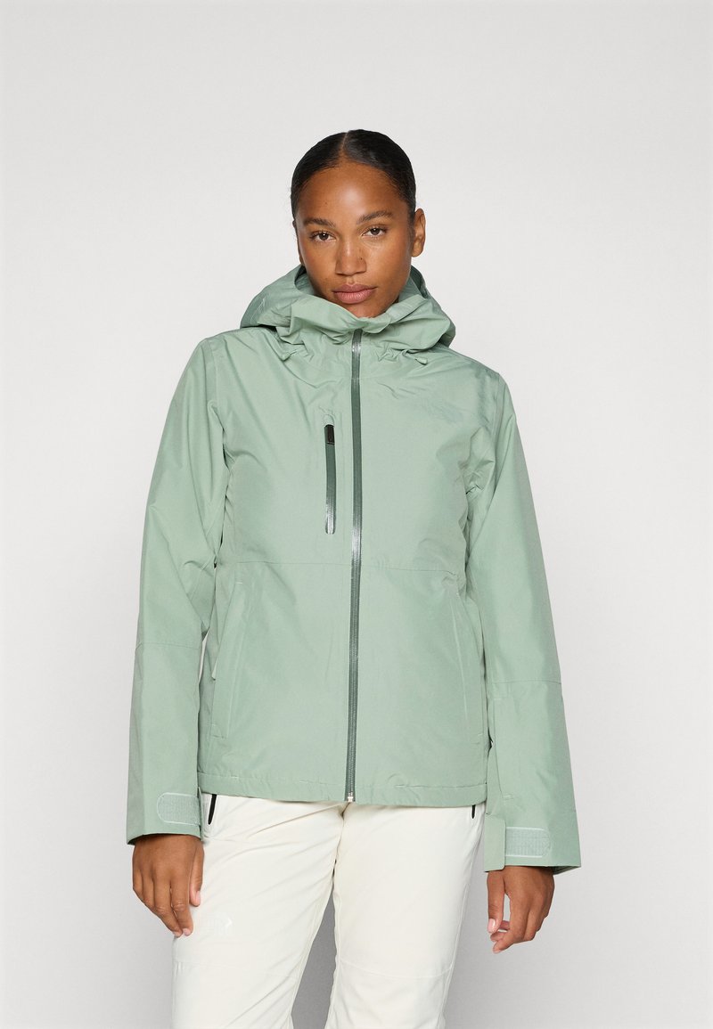 Chaqueta impermeable verde claro con capucha, que cuenta con un bolsillo en el pecho con cremallera y puños ajustables. Textura suave y diseño moderno.
