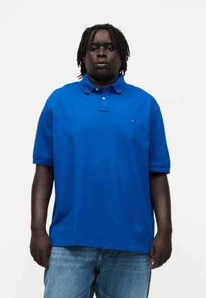 Uomo con dreadlocks che indossa una polo azzurro brillante a maniche corte e jeans azzurri chiari, in piedi davanti a uno sfondo bianco uniforme.
