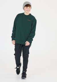 Grön sweatshirt med orange logga, i kombination med svarta tapered byxor och svarta träningsskor, bärs med en grå stickad mössa.
