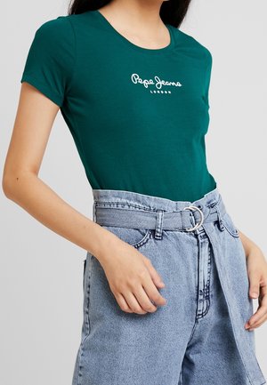 Kobieta ubrana w dopasowany, ciemnozielony t-shirt Pepe Jeans London oraz jasnoniebieskie jeansy z wysokim stanem z materiałowym paskiem i metalową klamrą.