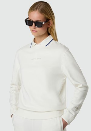 Femme portant un polo blanc à manches longues avec le texte « North Sails », mains dans les poches, et lunettes de soleil rectangulaires noires, sur un fond uni.