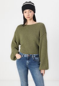 Olivgrön stickad tröja med vida ärmar och en lös passform, matchad med blå denimjeans. Modellen bär en svart stickad mössa med vita ränder.
