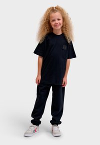 Svart kortärmad t-shirt med en fyrkantig lapp, kombinerad med lösa svarta byxor och vita sneakers med röda och blå detaljer.