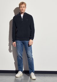 Pull-over en maille bleu marine à zip quart avec une texture côtelée, associé à un jean bleu clair et des baskets blanches. Présente un petit logo sur la poitrine.