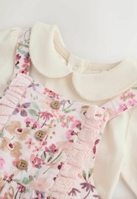 Chemise bébé couleur crème avec un col arrondi sous une robe salopette fleurie rose avec des volants et des boutons en bois.