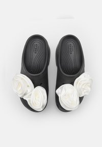 Black clogs with white satin rose accents on the front, featuring a textured insole and ventilation holes along the sides.
