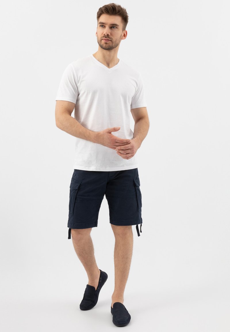 T-shirt bianca in cotone a V abbinata a pantaloni cargo blu marino con tasche laterali e cordini regolabili. Indossata con scarpe slip-on blu marino.