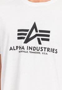 Λευκό βαμβακερό T-shirt με μαύρο γραφικό λογότυπο που περιλαμβάνει καλλιτεχνικά γράμματα «A» και ρίγες, με κείμενο της μάρκας και τοποθεσία από κάτω.