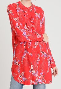 Blouse florale rouge à manches longues, avec un devant à volants et une fermeture à boutons. Présente des fleurs violettes et blanches sur un fond texturé.