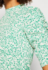 Blouse blanche à motif floral feuillu vert avec manches bouffantes froncées sur une personne aux cheveux bouclés.