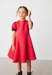 Punainen mekko, jossa on liehuva siluetti, puffihihat, joissa on mustia pitsikoristeita, teksturoitu kangas, jossa on hienovarainen kohokuvio.