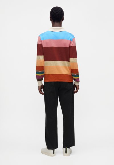 Homme debout, face à l'arrière, portant un pull rayé multicolore, un pantalon noir et des baskets blanches sur un fond clair uni.