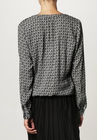 Blouse à manches longues à motifs noir et blanc avec un dos plissé et un col rond, fabriquée en tissu lisse. Présente des formes de diamant.