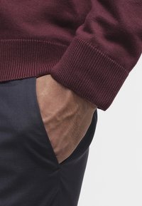 Mano destra nella tasca dei pantaloni scuri, indossa un maglione a maniche lunghe bordeaux con polsini a coste visibili.