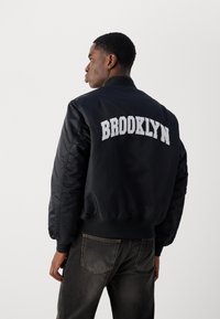 Crna bomber jakna sjajnog izgleda, s natpisom "BROOKLYN" izvezenim bijelom bojom na leđima, rebrastim manžetama i pojasom.