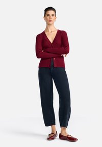 Cardigan côtelé bourgogne avec un décolleté en V profond et une fermeture à bouton, associé à un jean bleu foncé à coupe décontractée et des chaussures plates bourgogne.