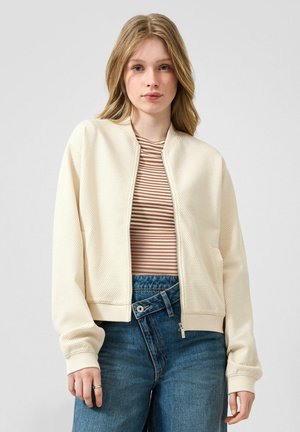 Giovane donna con capelli biondi indossa una giacca crema con zip e texture, una camicia a righe marrone e beige e jeans blu a vita alta con una particolare sovrapposizione di bottoni.