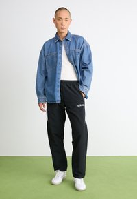Denim skjorte med knapper og to forlommer, parret med sorte træningsbukser med hvid Adidas-logo og blå sidelister, båret med hvide sneakers.
