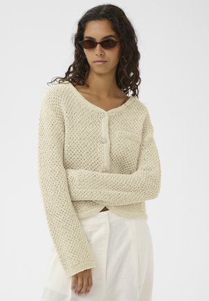 Femme portant des lunettes de soleil foncées et un cardigan beige en maille avec des boutons, associée à un pantalon blanc taille haute, debout les bras croisés.