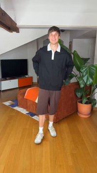 Νέος άνδρας με μαύρο μακρυμάνικο πουκάμισο, καφέ σορτς και άσπρα αθλητικά παπούτσια, στέκεται σε ένα σαλόνι με ένα μεγάλο φυτό και τηλεόραση στο παρασκήνιο.