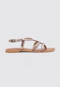 Sandalia plana de oro rosa con correas metálicas trenzadas, hebilla ajustable y suela plana de color marrón, diseñada para la comodidad y facilidad de uso.
