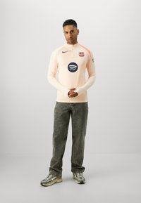 Light peach long-sleeve jersey with a zip neck, ribbed texture, Barcelona logo, and a central blue Spotify logo. Worn with gray jeans and sneakers.