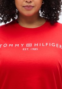 Κόκκινο βαμβακερό T-shirt με λευκή επιγραφή "TOMMY HILFIGER" και κάτω από αυτήν το "EST. 1985". Μανίκια κοντά, κλασικός στρογγυλός γιακάς, απαλό ύφασμα.