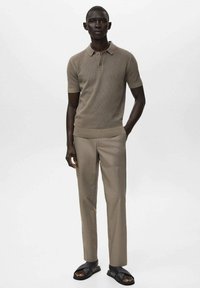 Polo shirt lavorato a maglia color tan con maniche corte, abbinato a pantaloni beige chiaro e sandali neri. Il tessuto ha una finitura texturizzata.