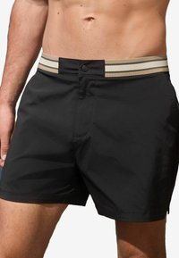 Shorts de bain noirs en tissu léger, dotés d'une ceinture élastique rayée beige et noire et d'une fermeture à bouton à l'avant.