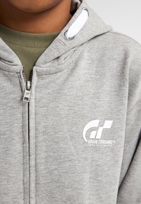 Felpa grigia con zip realizzata in tessuto morbido, caratterizzata da un logo "Gran Turismo" bianco e un cordino bianco a contrasto sulla cappuccio.