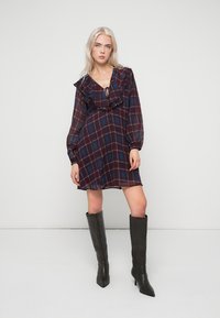 Ριγέ φόρεμα σε κόκκινο και ναυτικό μπλε χρώμα με ρέον ντεκολτέ, μακριά μανίκια και χαλαρή εφαρμογή. Το μοντέλο φοράει μαύρες μπότες μέχρι το γόνατο.