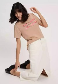 Beżowy T-shirt z wyraźnym różowym napisem, zestawiony z białą spódnicą do kolan z bocznymi rozcięciami oraz czarnymi butami na platformie; styl casual.