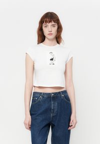 Witte cropped t-shirt met korte mouwen, voorzien van een zwart-wit grafisch print op de voorkant. Gecombineerd met donkerblauwe high-waisted jeans.