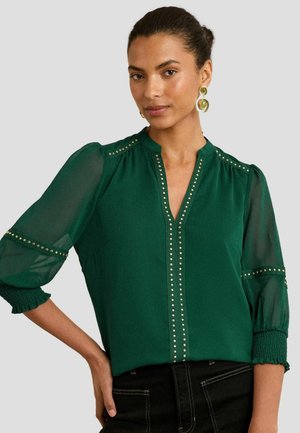 Blusa verde scuro con maniche trasparenti a 3/4, scollo a V e dettagli in borchie metalliche lungo il davanti e le maniche. I polsini arricciati aggiungono texture.