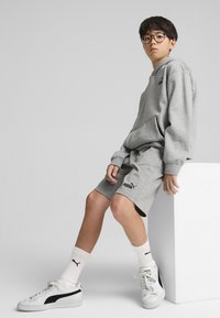 Un sweat à capuche gris et un short en tissu doux, associés à des chaussettes blanches et des baskets Puma avec des accents noirs. Le sujet est assis sur un bloc blanc.