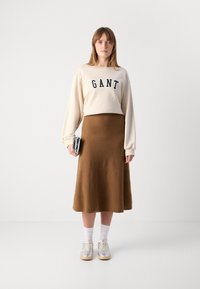 Kvinna står klädd i en krämfärgad sweatshirt med texten "GANT", brun midiklänning, vita strumpor, silverfärgade sneakers och håller en metallisk clutchväska.