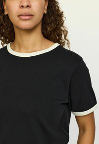 T-shirt noir à manches courtes avec un col et des ourlets de manches en crème contrastants, fabriqué en tissu coton doux. Texture lisse, coupe décontractée.