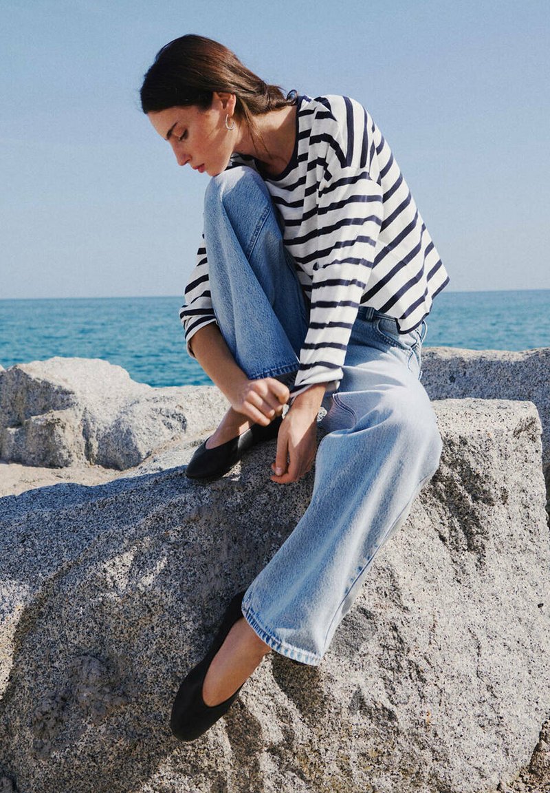 Femme portant une chemise rayée et un jean, assise sur un grand rocher au bord de la mer, ajustant des chaussures noires sous un ciel bleu clair.