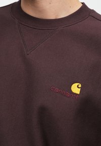 Felpa color marrone con scollo rotondo a costine e piccolo logo ricamato in giallo e rosso sul petto. Tessuto morbido e strutturato.