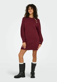 Robe pull tricotée bordeaux avec des manches surdimensionnées, présentant un ourlet côtelé et trois boutons décoratifs sur l'épaule gauche.
