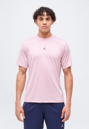 Jeune homme aux cheveux bouclés portant un t-shirt de sport rose clair et un short bleu marine, debout devant un fond clair uni.