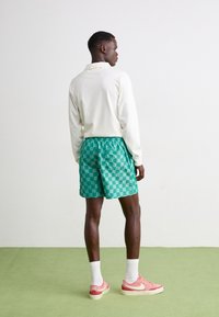 Grønne sjakkmønstret shorts laget av lett stoff, sammen med en hvit langermet skjorte og rosa sneakers, står på grønt gulv.