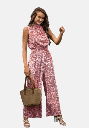 Lachende vrouw die een rood-wit bloemenmouwloze jumpsuit draagt, witte sandalen met hak en een gevlochten strohandtas vasthoudt.