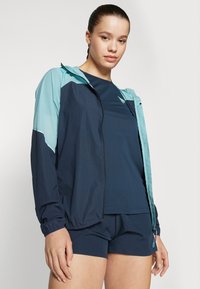 Marineblå og teal vindjakke med lett design, frontglidelås og elastiske mansjetter, kombinert med en matchende marineblå topp og shorts.