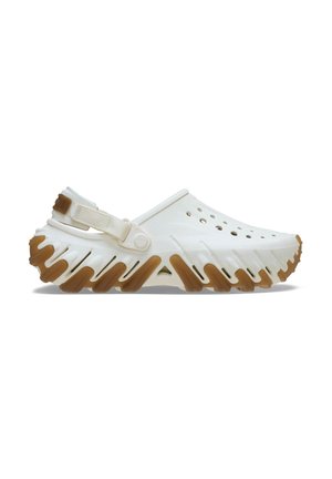 Sabots en plastique blanc avec un design à bout ouvert, des trous de ventilation sur les côtés et une semelle en caoutchouc texturée avec un motif en zigzag et des accents marron.